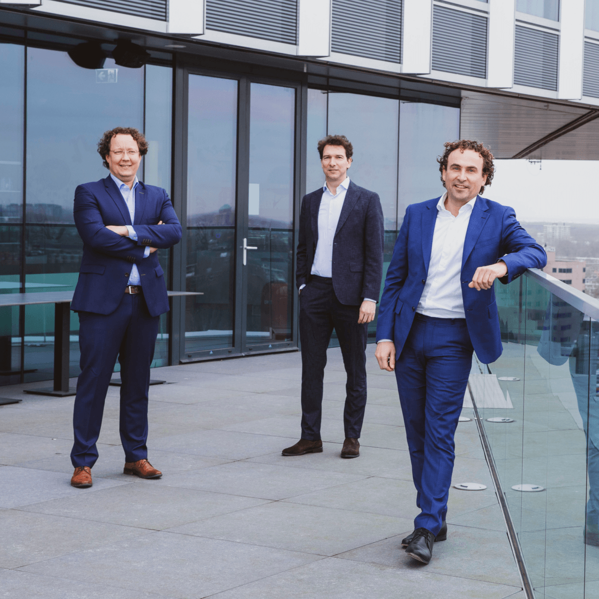 Three HVK Stevens advisors in suits on a modern office terrace with glass railing and city view